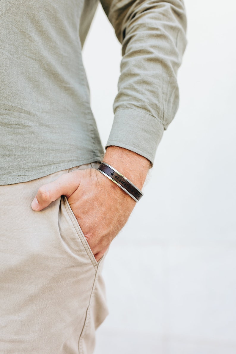 May include: A brown leather bracelet with a silver band and engraved coordinates. The bracelet is worn on a person's wrist.