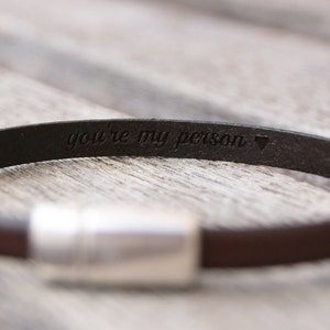 May include: A brown leather bracelet with a silver clasp. The bracelet is engraved with the words "you're my person" and a heart symbol.