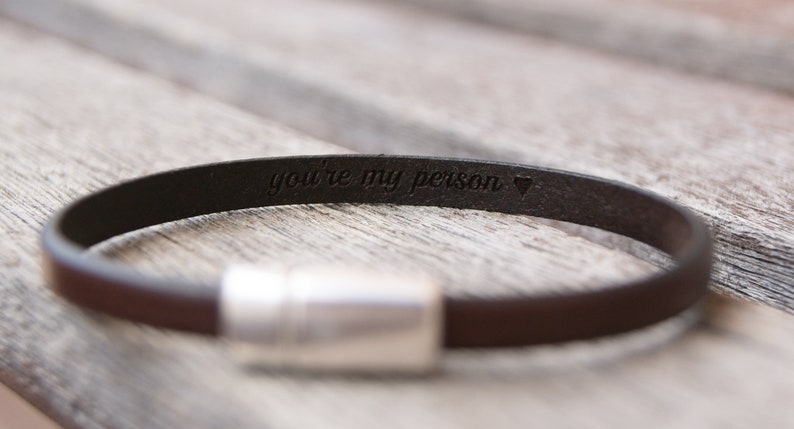 May include: A brown leather bracelet with a silver clasp. The bracelet is engraved with the words "you're my person" and a heart symbol.