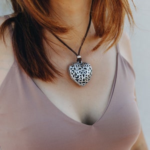 May include: A silver heart-shaped pendant necklace with a delicate filigree design hangs on a brown leather cord. The necklace is worn by a person with brown hair and a light brown tank top.