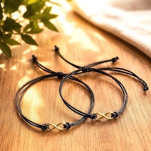Può includere: Due braccialetti neri con simboli dell'infinito color oro. I braccialetti sono su una superficie di legno, con uno sfondo sfocato di foglie verdi e un panno beige. I braccialetti sono legati con nodi regolabili.