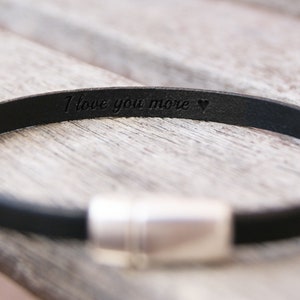 May include: A black leather bracelet with a silver clasp. The bracelet is engraved with the words "I love you more" and a heart.