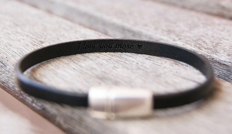 May include: A black leather bracelet with a silver clasp. The bracelet is engraved with the words "I love you more" and a heart.