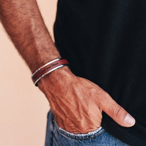 May include: A silver and burgundy cuff bracelet with the words "Brave - Loyal and Fierce" engraved on the burgundy band. The bracelet is worn on a wrist, with a black shirt and blue jeans in the background.