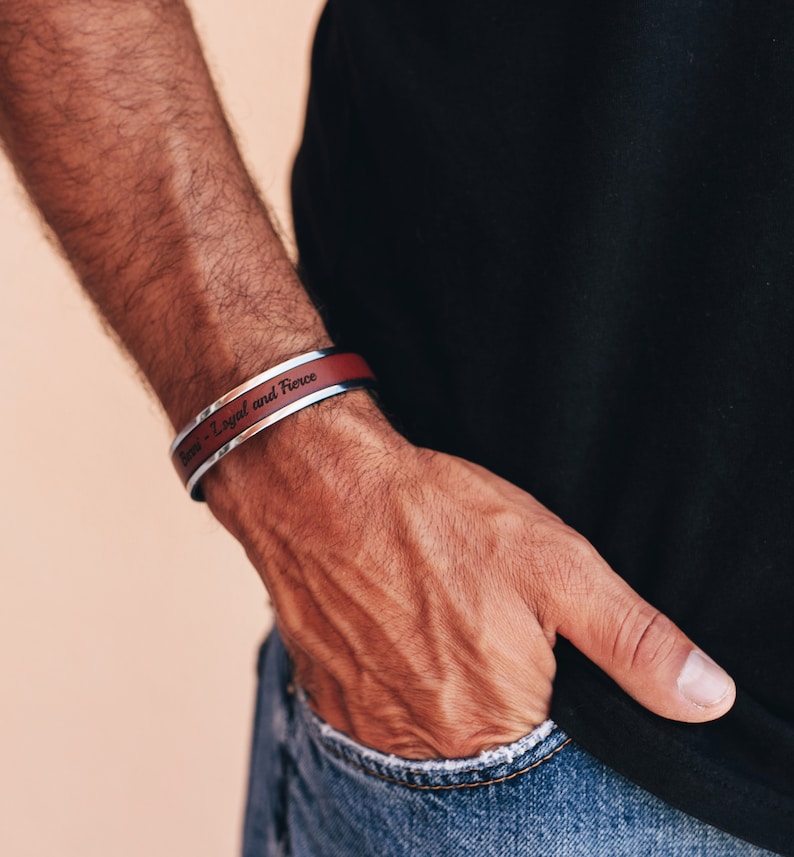 May include: A silver and burgundy cuff bracelet with the words "Brave - Loyal and Fierce" engraved on the burgundy band. The bracelet is worn on a wrist, with a black shirt and blue jeans in the background.