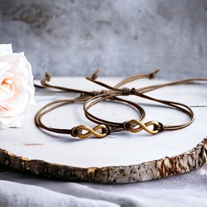 May include: Two matching infinity symbol bracelets. Each bracelet features a gold-colored infinity charm on a brown cord. The bracelets are adjustable and tied with a knot. The background includes a white flower and a wooden surface.