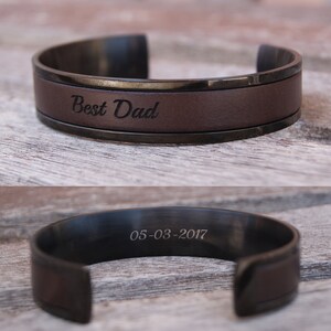 May include: A brown leather cuff bracelet with a black metal band. The bracelet is engraved with the words "Best Dad" and the date "05-03-2017".