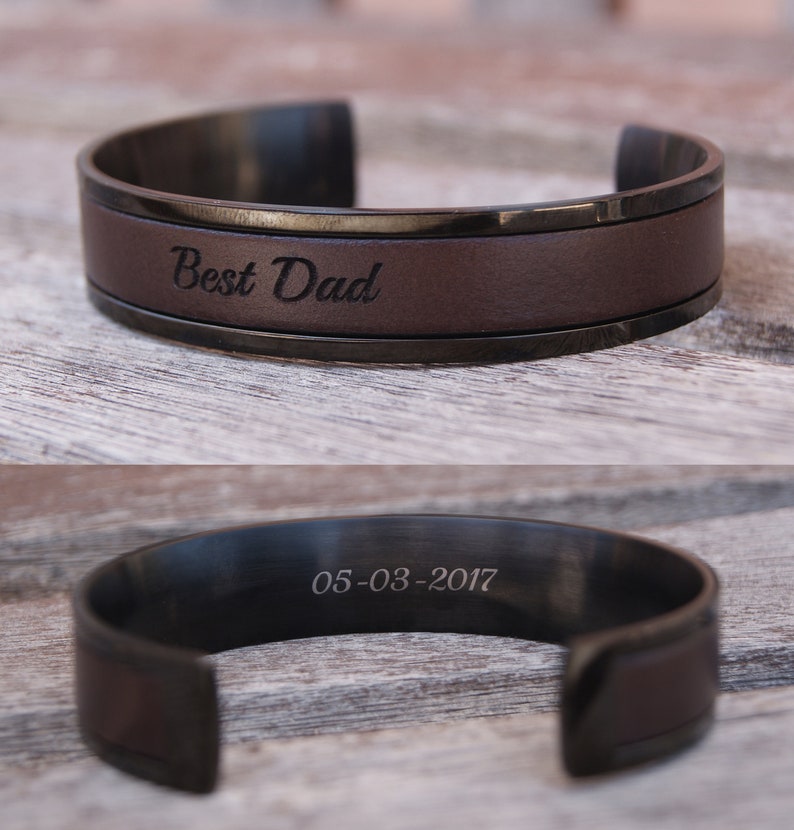 May include: A brown leather cuff bracelet with a black metal band. The bracelet is engraved with the words "Best Dad" and the date "05-03-2017".