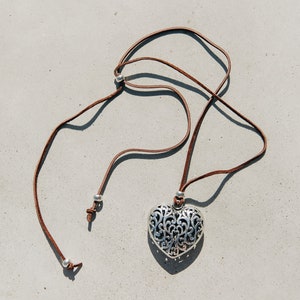 May include: A silver heart-shaped pendant with an intricate design hangs from a brown leather cord. The cord has silver beads at the ends.