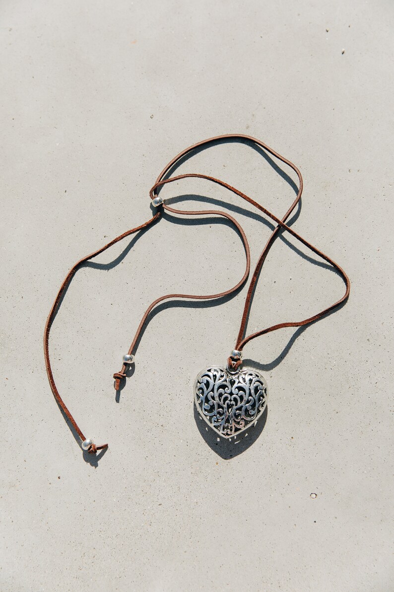 May include: A silver heart-shaped pendant with an intricate design hangs from a brown leather cord. The cord has silver beads at the ends.