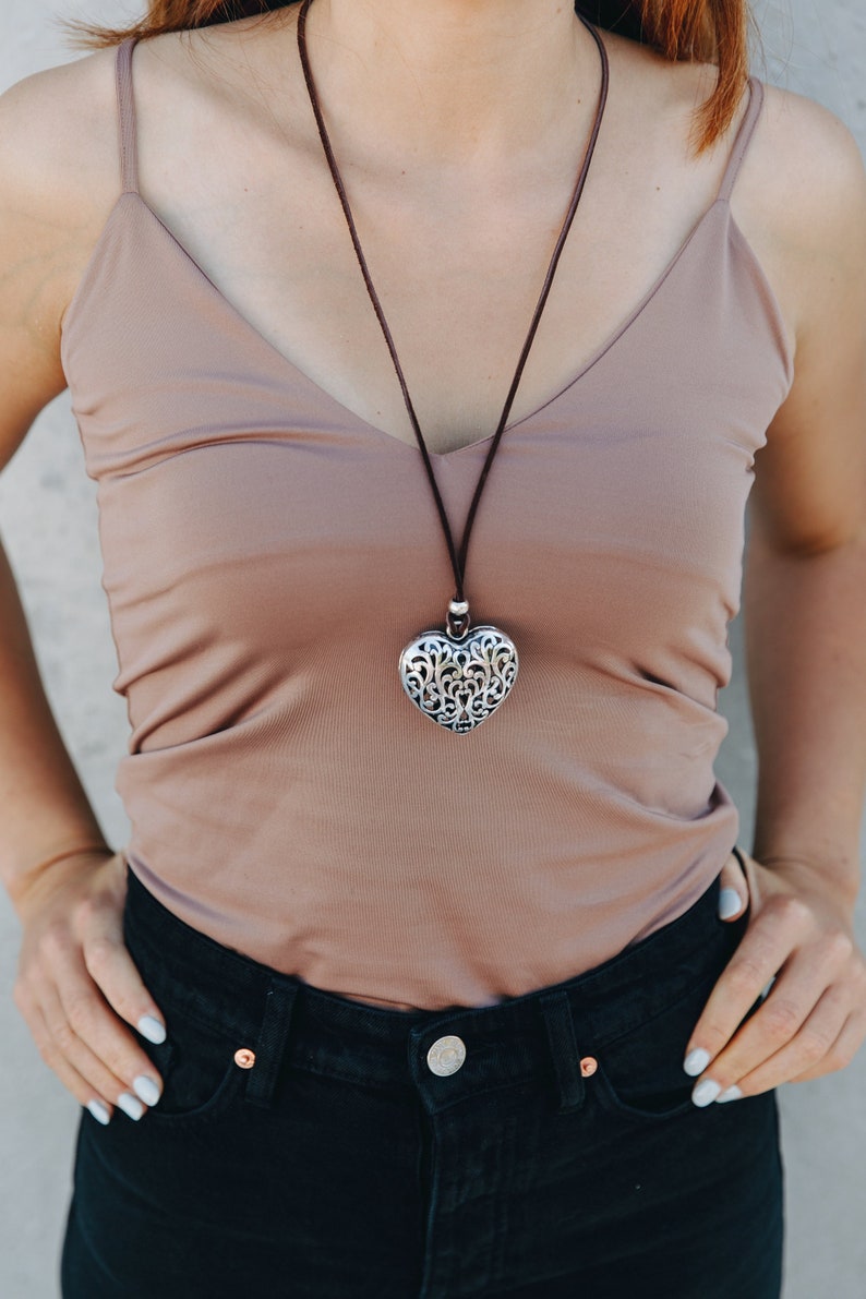 May include: A woman wearing a brown tank top and black jeans with a silver heart pendant necklace. The heart pendant has a detailed filigree design.