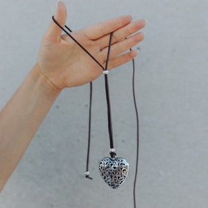 May include: A silver heart-shaped pendant with intricate filigree details hangs from a brown leather cord. The cord has silver beads and is tied in a knot at the end.