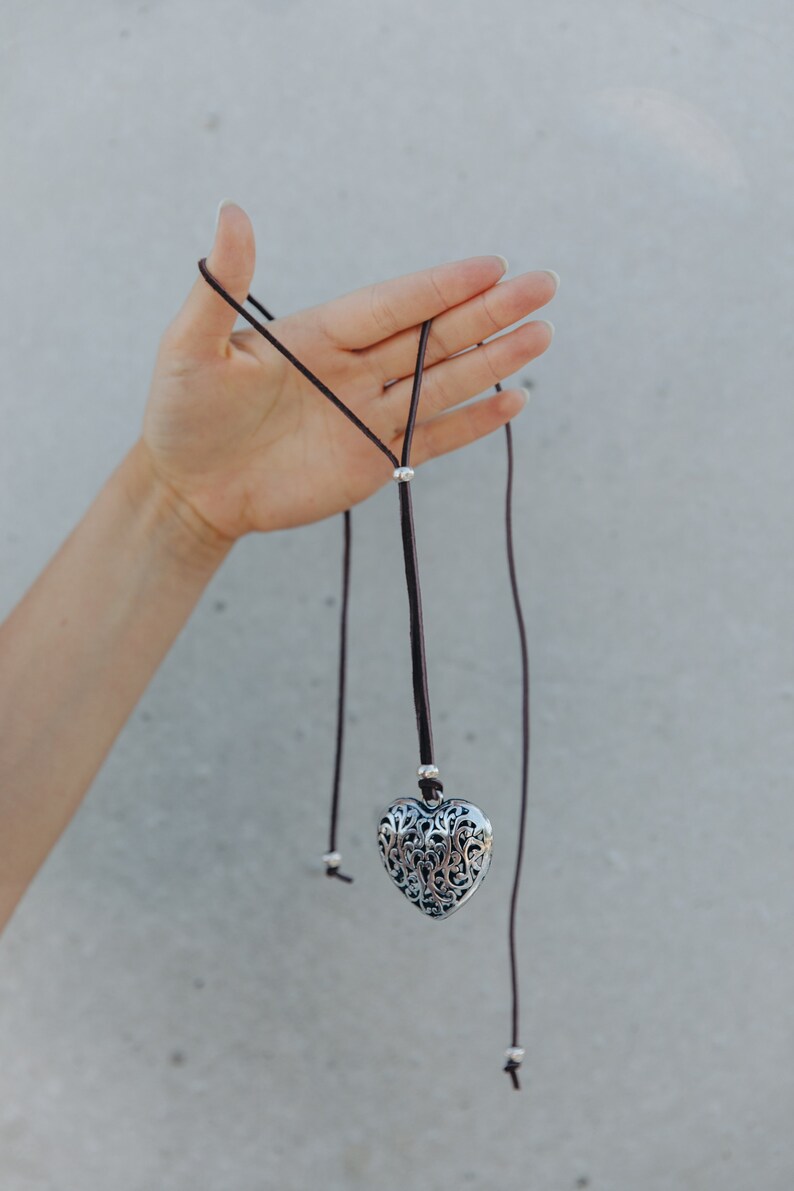 May include: A silver heart-shaped pendant with intricate filigree details hangs from a brown leather cord. The cord has silver beads and is tied in a knot at the end.