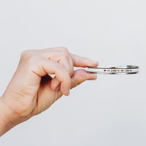 May include: A silver bracelet with a white band and a red star with the coordinates "41.2486 N, 96.1386 W" engraved on it.