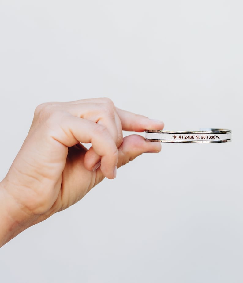 May include: A silver bracelet with a white band and a red star with the coordinates "41.2486 N, 96.1386 W" engraved on it.