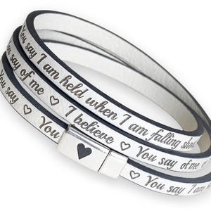 May include: White wrap bracelet with black trim and a silver clasp. The bracelet has engraved text and heart symbols. The text reads "You say I am held when I am falling" and "You say of me I believe".
