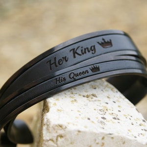 Op de afbeelding: Een paar zwarte metalen armbanden met de woorden "Her King" en "His Queen" gegraveerd, elk met een klein kroonsymbool.
