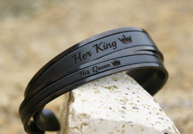 Op de afbeelding: Een paar zwarte metalen armbanden met de woorden "Her King" en "His Queen" gegraveerd, elk met een klein kroonsymbool.