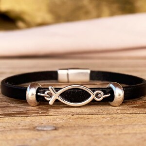 Könnte beinhalten: Schwarzes Lederarmband mit einem silbernen Ichthys-Fischsymbol. Das Armband hat einen Magnetverschluss und silberne Akzente. Präsentiert auf einer Holzoberfläche.