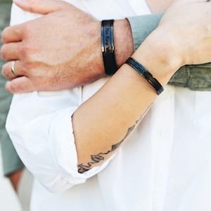Op de afbeelding: Een stel draagt bijpassende zwarte armbanden met de woorden "His King" en "Her Queen" erop gegraveerd. De vrouw draagt een wit shirt en heeft een tatoeage op haar arm.
