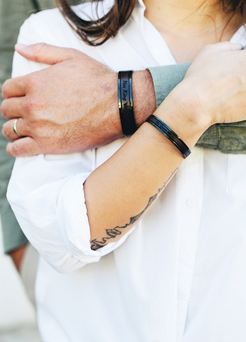 Op de afbeelding: Een stel draagt bijpassende zwarte armbanden met de woorden "His King" en "Her Queen" erop gegraveerd. De vrouw draagt een wit shirt en heeft een tatoeage op haar arm.