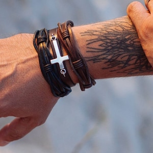 May include: A man's wrist with a leather bracelet featuring a silver cross charm. The bracelet is made of multiple strands of brown leather and has a braided design. The man's arm also has a black tattoo of a tree branch.