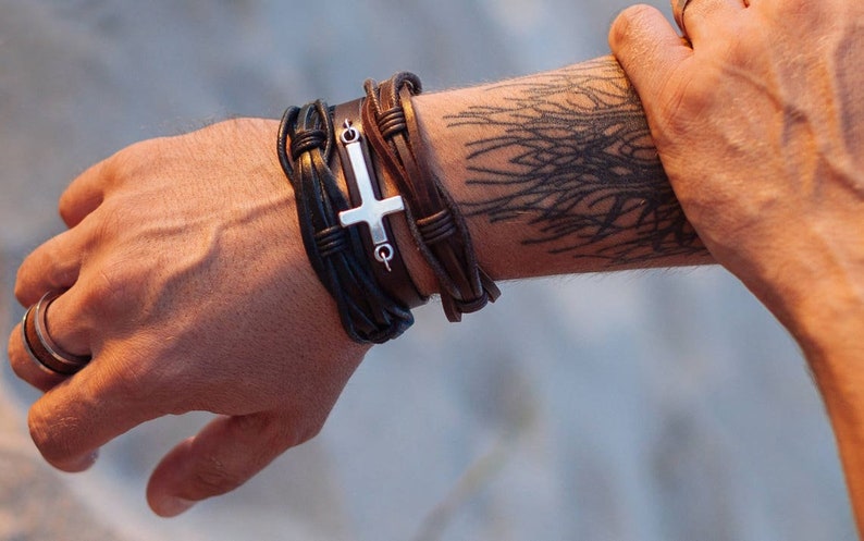 May include: A man's wrist with a leather bracelet featuring a silver cross charm. The bracelet is made of multiple strands of brown leather and has a braided design. The man's arm also has a black tattoo of a tree branch.