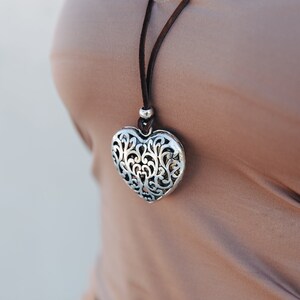 May include: A silver heart-shaped pendant necklace with a delicate filigree design hangs on a brown leather cord.
