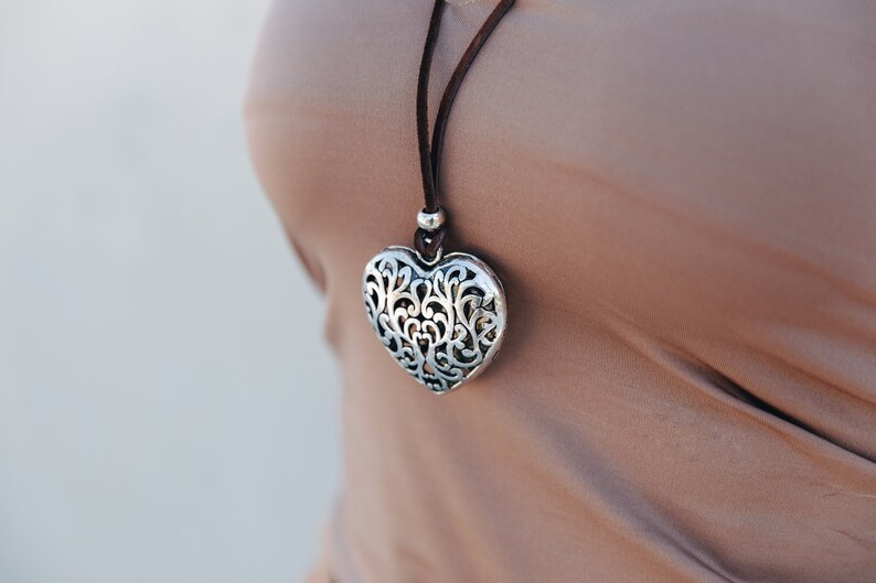 May include: A silver heart-shaped pendant necklace with a delicate filigree design hangs on a brown leather cord.