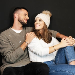 May include: A couple sitting together, the woman is wearing a white knit hat and a white sweater, the man is wearing a gray sweater. They are both looking at each other and smiling.