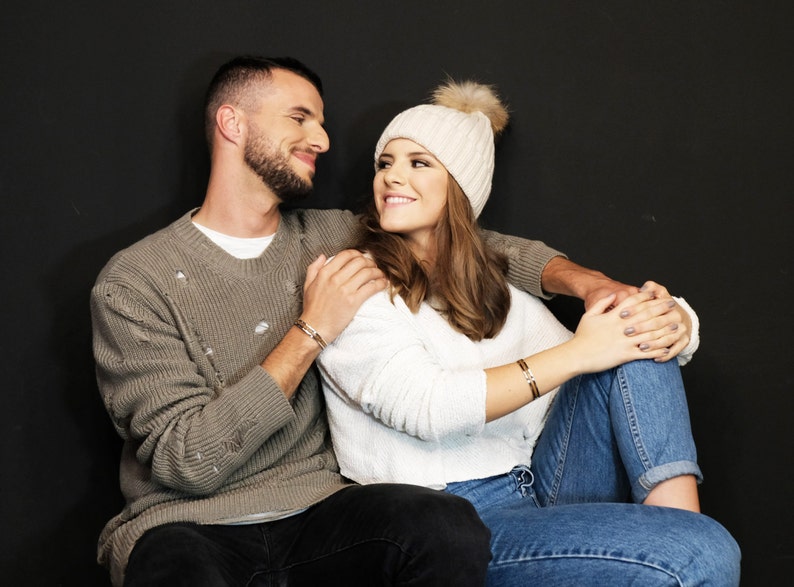 May include: A couple sitting together, the woman is wearing a white knit hat and a white sweater, the man is wearing a gray sweater. They are both looking at each other and smiling.
