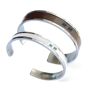 Puede incluir: Dos pulseras de puño de acero inoxidable. Una tiene una incrustación de cuero marrón, la otra blanca. Ambas pulseras tienen marcas de conteo grabadas. Las pulseras se muestran sobre un fondo blanco.