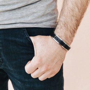 May include: A black leather cuff bracelet with a silver band engraved with the coordinates 46.58.21.7. The bracelet is worn on a person's wrist.