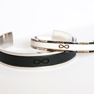 Puede incluir: Dos pulseras a juego con un símbolo de infinito. Una pulsera tiene una banda de cuero negro y la otra tiene una banda blanca. Ambas pulseras están hechas de acero inoxidable.