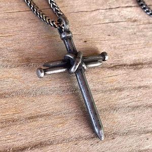 Może przedstawiać: Ciemny metalowy wisiorek w kształcie krzyża wykonany z dw&oacute;ch skrzyżowanych gwoździ, posiadający zwietrzałe, oksydowane wykończenie. Krzyż wisi na pasującym ciemnosrebrnym łańcuszku o splocie kłosowym, prezentując surowy, industrialny wygląd.