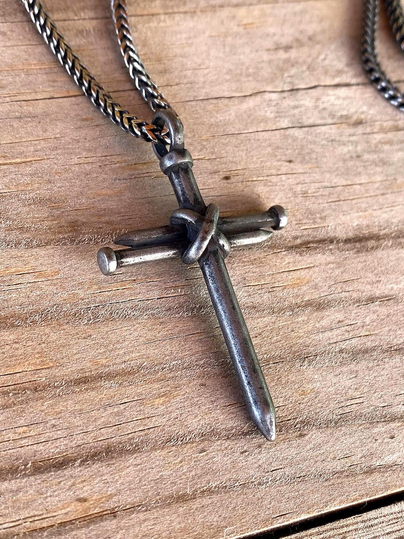 Może przedstawiać: Ciemny metalowy wisiorek w kształcie krzyża wykonany z dw&oacute;ch skrzyżowanych gwoździ, posiadający zwietrzałe, oksydowane wykończenie. Krzyż wisi na pasującym ciemnosrebrnym łańcuszku o splocie kłosowym, prezentując surowy, industrialny wygląd.