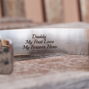 May include: Silver cuff bracelet with a brown leather band. The bracelet is engraved with the words "Daddy, My First Love, My Forever Hero."