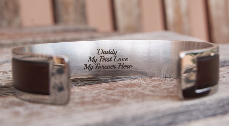 May include: Silver cuff bracelet with a brown leather band. The bracelet is engraved with the words "Daddy, My First Love, My Forever Hero."