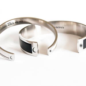 May include: Two silver bangle bracelets with black accents. One bracelet has the inscription "This is us" and a crescent moon symbol. The other bracelet has the inscription "forever" and a sun symbol.