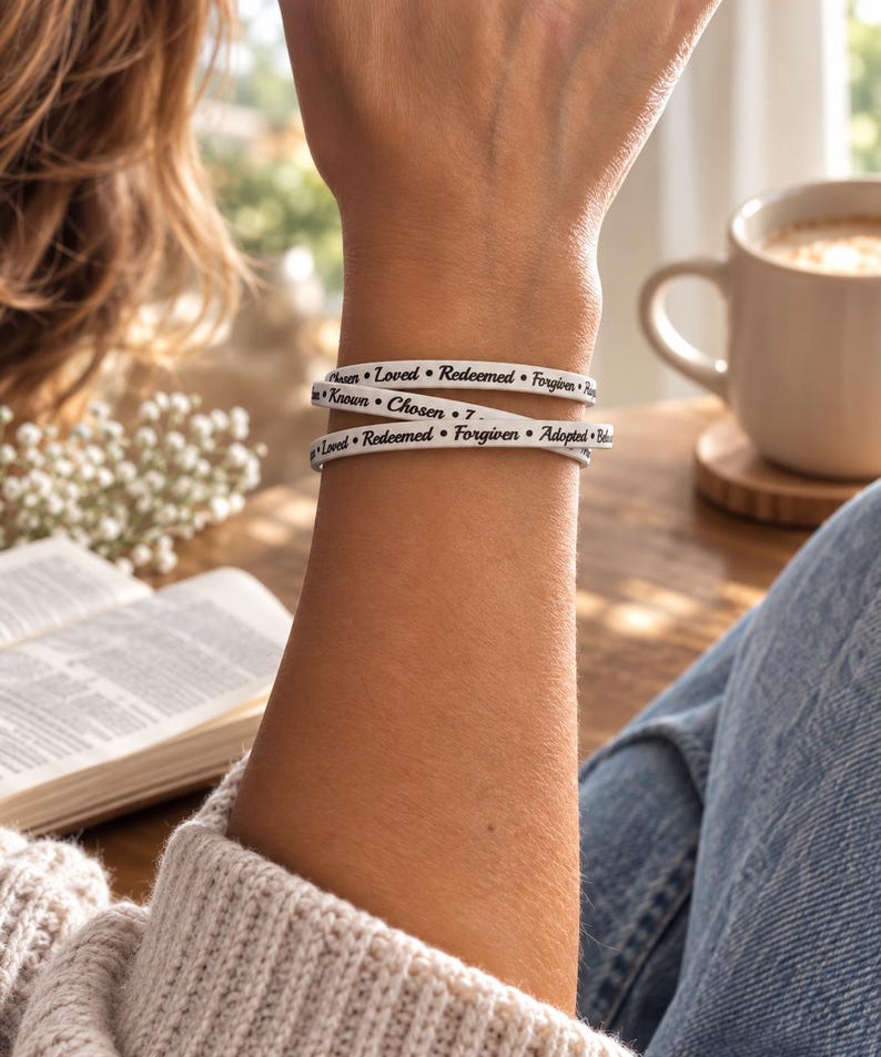 Puede incluir: Una pulsera envolvente blanca con texto negro impreso en un patr&oacute;n repetitivo. Las palabras visibles incluyen Loved, Redeemed, Forgiven, Known, Chosen y Adopted, separadas por peque&ntilde;os puntos negros. La pulsera rodea la mu&ntilde;eca varias veces.
