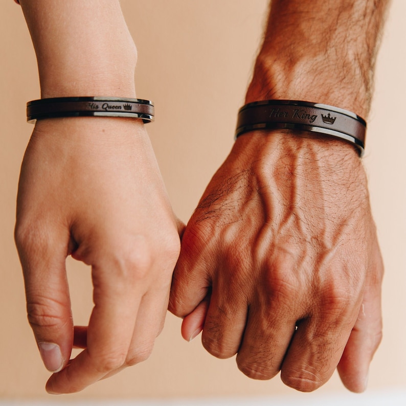 May include: Two people holding hands, each wearing a black leather bracelet with a silver inscription. The bracelet on the left hand says "His Queen" and the bracelet on the right hand says "His King".