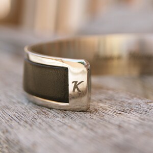May include: A silver cuff bracelet with a black leather band and the letter "K" engraved on the metal.
