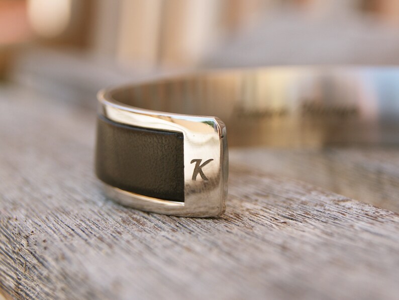 May include: A silver cuff bracelet with a black leather band and the letter "K" engraved on the metal.