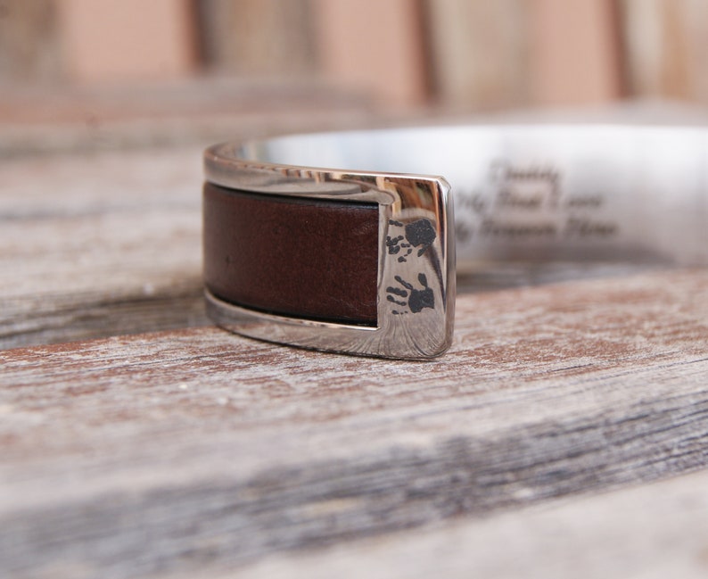 May include: A silver cuff bracelet with a brown leather band and engraved handprints. The bracelet is on a wooden surface.