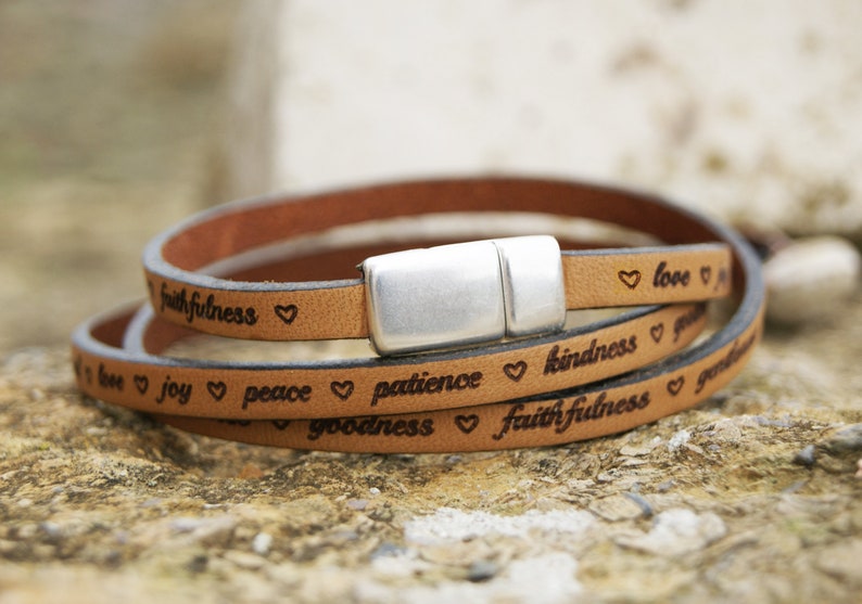 Puede incluir: Un brazalete de cuero marr&oacute;n con cierre de plata. El brazalete tiene las palabras "love", "joy", "peace", "patience", "kindness", "goodness" y "faithfulness" grabadas.