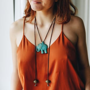Peut inclure: Un collier avec pendentif éléphant turquoise, long cordon marron et perles métalliques décoratives. Le pendentif est un éléphant stylisé et détaillé. Le collier est porté sur un caraco orange.