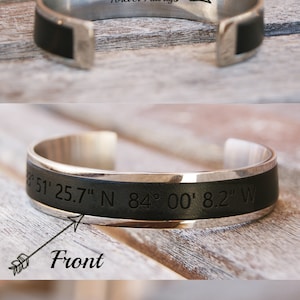 May include: A silver bracelet with a black leather band. The bracelet can be engraved with text on the inside, front, or ends. The image shows examples of engraving: "Forever Always", "51' 25.7" N 84&deg; 00' 8.2" W", and "K".
