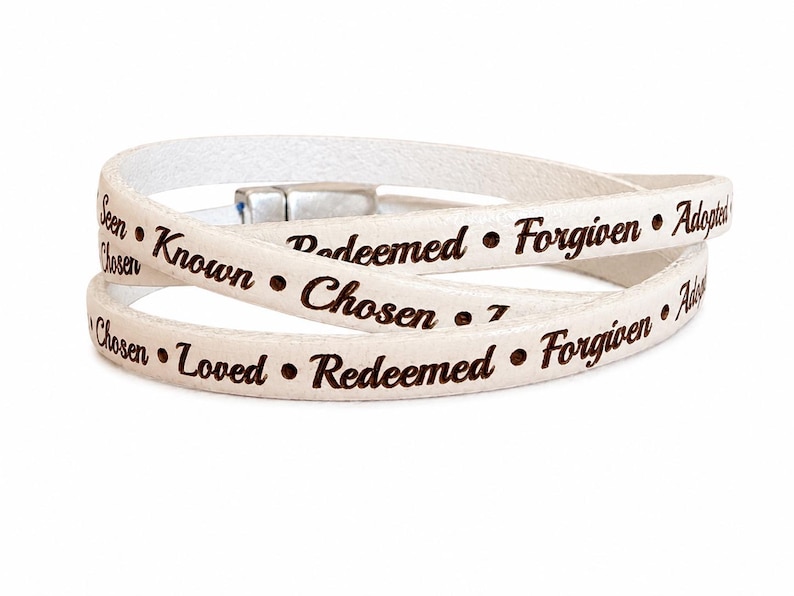 Puede incluir: Una pulsera envolvente blanca con texto grabado en marr&oacute;n oscuro que incluye las palabras Seen, Known, Redeemed, Forgiven, Adopted, Chosen y Loved, separadas por peque&ntilde;os puntos. La pulsera tiene un cierre magn&eacute;tico de metal plateado.