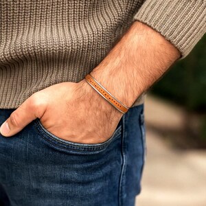 Könnte beinhalten: Ein braunes Lederarmband mit silbernem Rand und gepunktetem Design. Das Armband wird am Handgelenk getragen, kombiniert mit einem braunen Pullover und einer blauen Jeans. Ein stilvolles Accessoire.