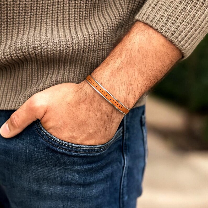 K&ouml;nnte beinhalten: Ein braunes Lederarmband mit silbernem Rand und gepunktetem Design. Das Armband wird am Handgelenk getragen, kombiniert mit einem braunen Pullover und einer blauen Jeans. Ein stilvolles Accessoire.